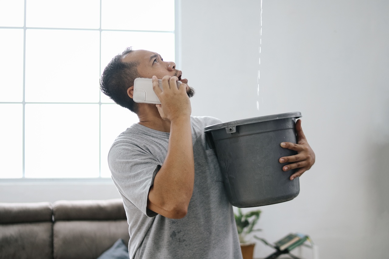 homeowner holding a bucket under a leaking ceiling while calling for plumbing help